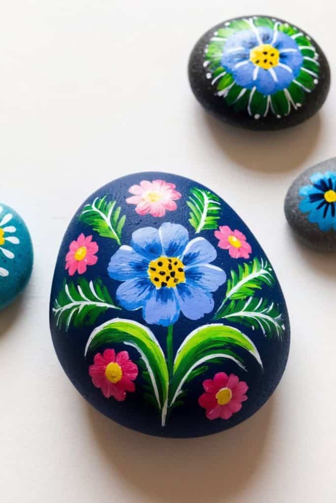 Painted stones featuring colorful flowers and leaves on a white surface. The largest stone shows a large blue flower surrounded by pink blossoms.