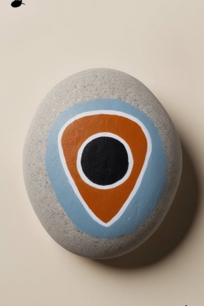 A smooth grey stone featuring concentric shapes painted in black, white, orange, and blue on a neutral background.