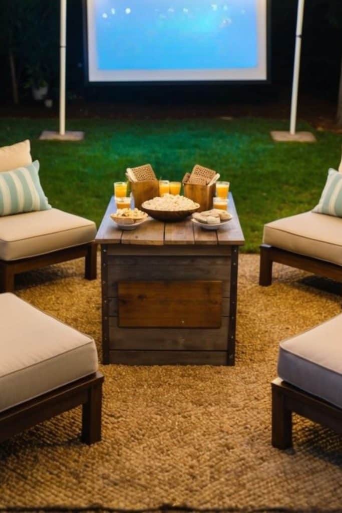 Outdoor seating arrangement with four cushioned chairs around a wooden table holding snacks and drinks. A projector screen is visible in the background on a lawn.