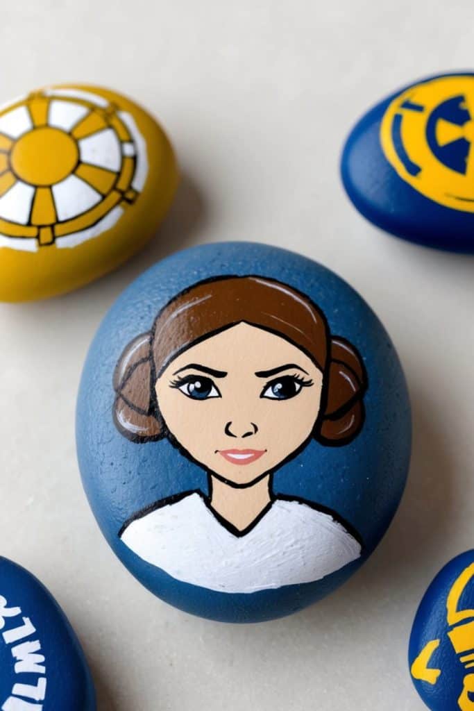 A painted rock features a cartoon depiction of Princess Leia with brown hair buns and a white outfit, surrounded by other painted rocks with yellow and blue designs.