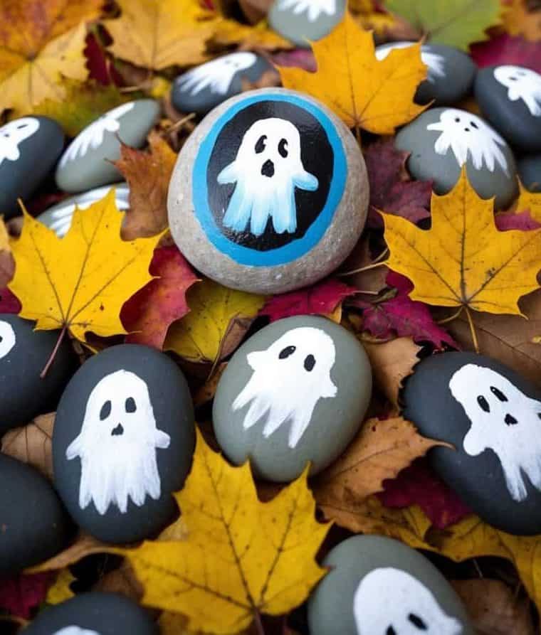 Painted rocks with ghost designs are scattered on autumn leaves, with illustrated ghosts appearing to float above the scene.