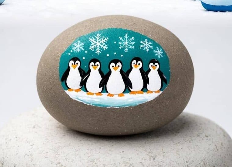 A group of penguins stands in snow with decorated eggs beside them; below, a stone is painted with five penguins on a snowy background.