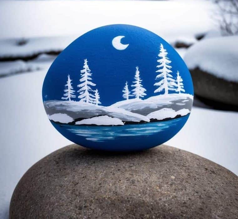 A painted rock with a winter scene sits on a stone in front of a snow-covered cabin surrounded by pine trees under a starry night sky.