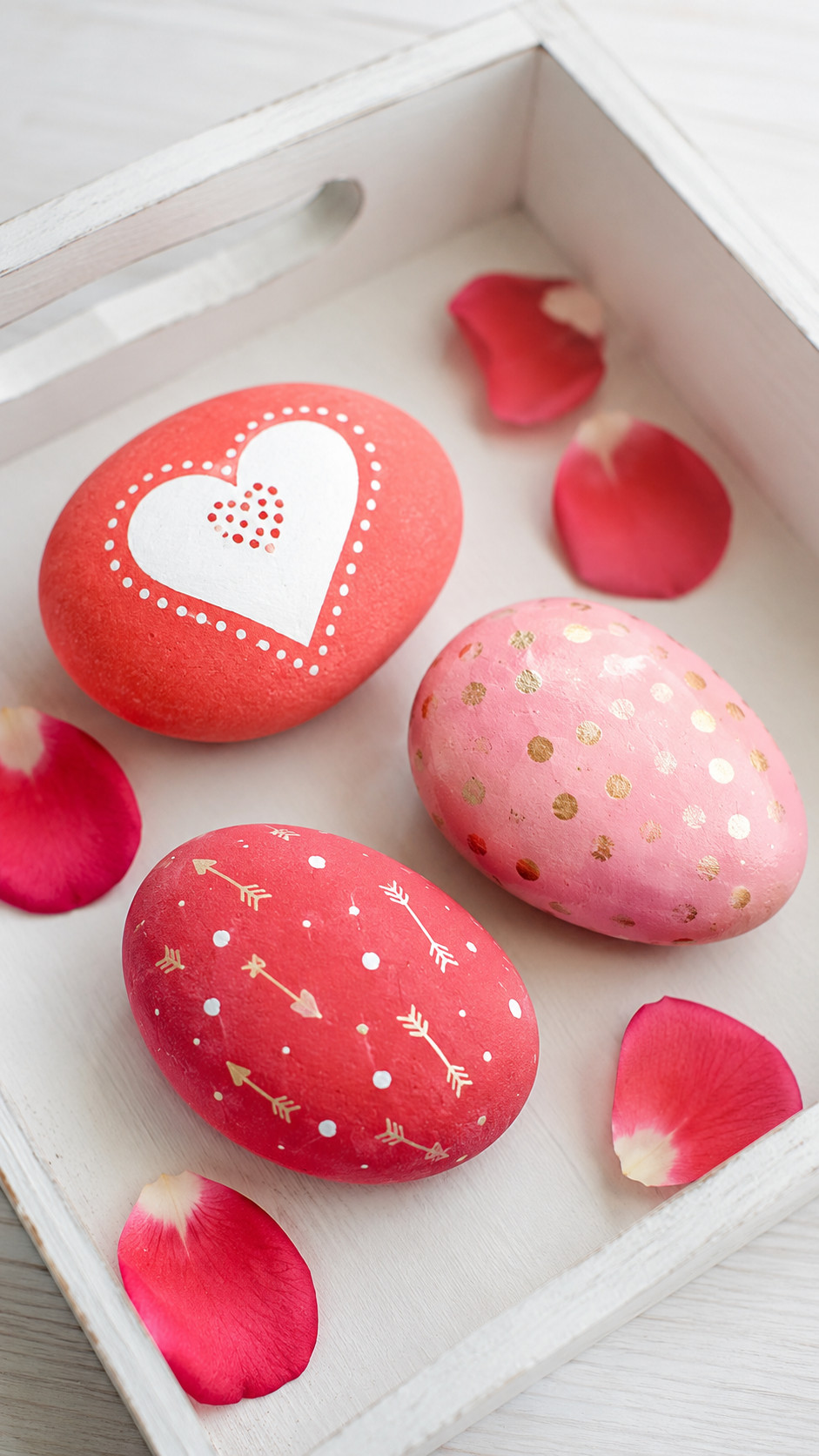 Photo of a marbled heart painted rock with swirls of pink, white, and metallic gold sitting on a wooden surface with rose petals nearby.