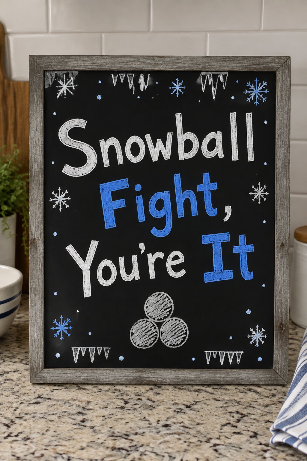 Snowball Fight Board: a framed winter chalkboard sign reading "Snowball Fight, You're It", hand-drawn in chalk with three round snowballs stacked in a small pyramid.